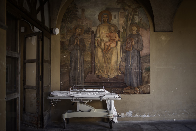A stretcher under a fresco in the courtyard of the Pesanti Fenaroli Hospital of Alzano Lombardo (Bergamo) found itself at the center of the Covid-19 outbreak, Italy on March 16, 2020. In March Italy found itself at the center of the Covid-19 epidemic, and its northern province of Bergamo become the ground zero for the coronavirus in all the world. In a few weeks, the number of infections and casualties have reached the highest levels worldwide heralding a global crisis that would soon spread in all the other countries. The public hospital system, particularly the ICU were collapsing due to the high number of infected cases and seriously ill patients, many of them dying at home with no chance to been hospitalized. The Italian army has organized operations to transport dozens of coffins of Covid-19 deceased to other regions for cremation because local morgues were full. In two months the official number of deaths in Italy is more than 32.000, of which 15.296 only in Lombardy.